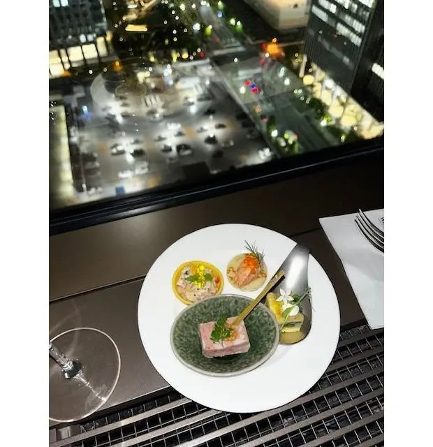 高層階の窓から夜景を望む様子。テーブルには白い皿に盛り付けられた、4つの小さな料理（魚介のタルト、ピンク色のゼリー、エビのパイ、白身魚のムース）が置かれている。左にはグラス、右にはメニューとナプキンがある。