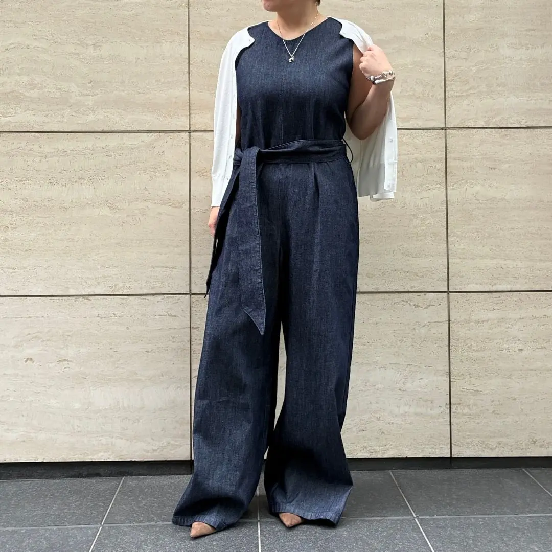 濃い青のデニム素材のワイドレッグパンツとトップスが一体になったジャンプスーツ。腰に黒いベルトがあり、白いボタン付きのカーディガンを羽織っている。女性が全身を写した正面からの写真。