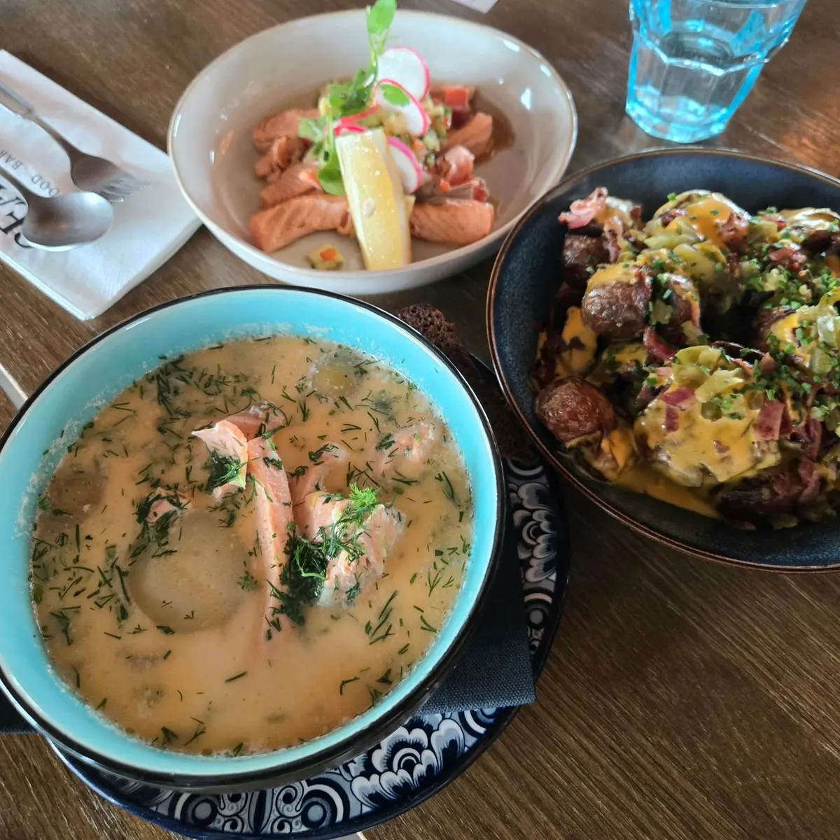 テーブルに置かれた3つの料理。左は淡いクリーム色のスープで、赤い魚と緑のハーブが浮かんでいる。中央は赤と黄色の野菜サラダ。右は、肉とジャガイモが混ざった盛り付けられた料理。青いグラスが近くに置かれている。