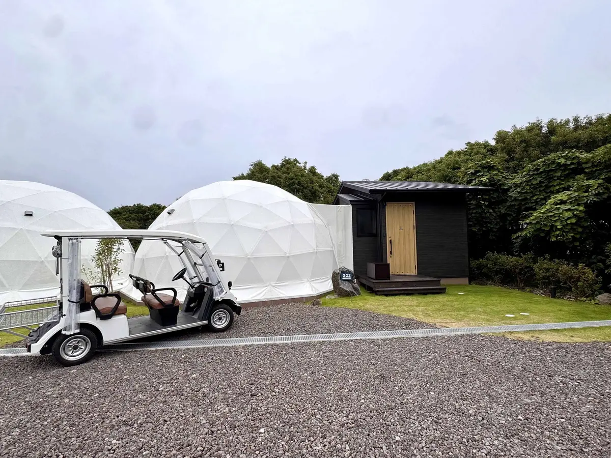 白い電動カートが、丸い白いドーム型テントと黒い建物の前で止まっている。カートには茶色の座席があり、建物の入り口には木製のドアがある。背景には緑の木々が広がっている。