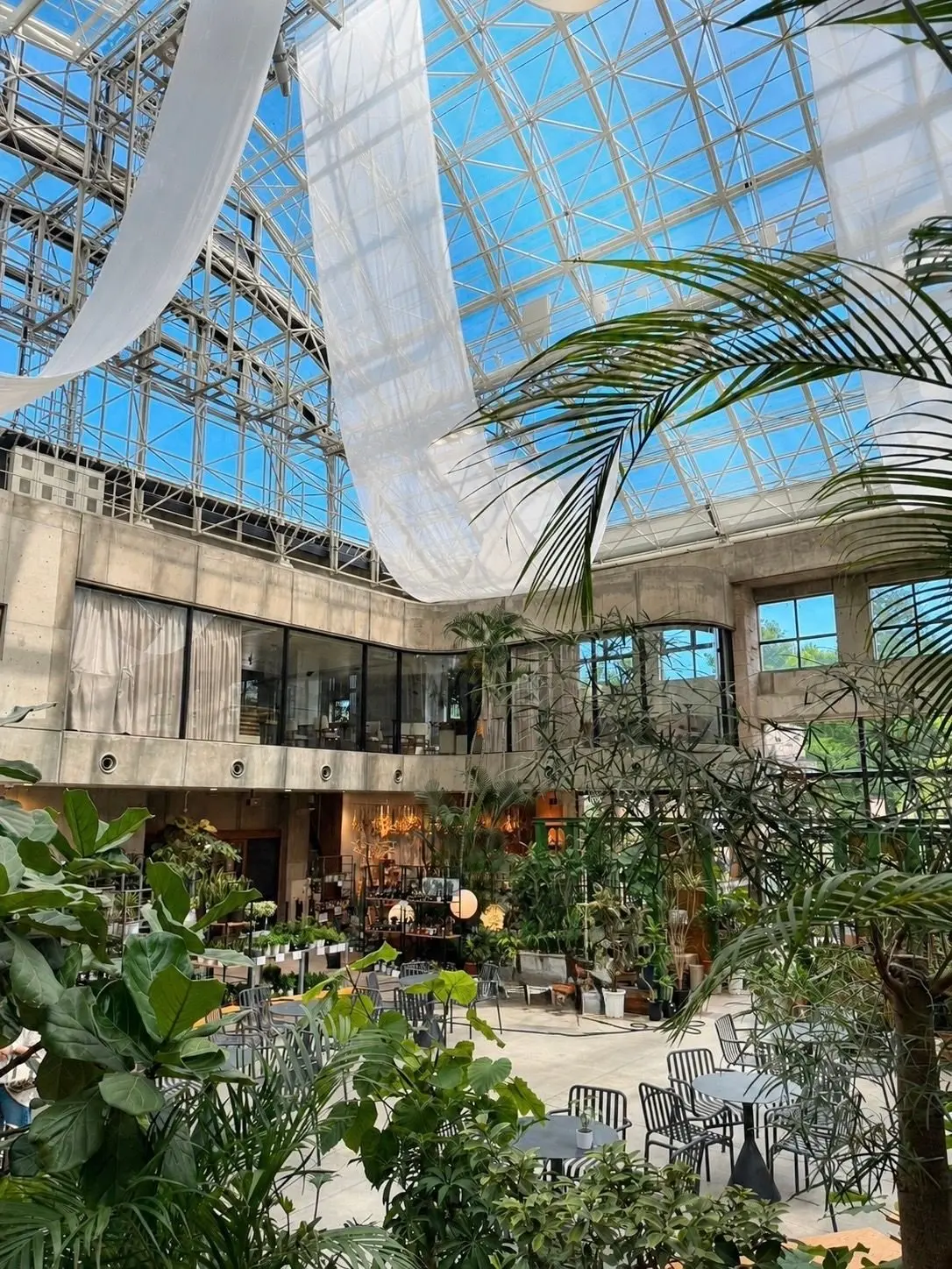 ガラスの天井付きの屋内植物園で、ヤシや他の緑豊かな植物が広がり、テラス席とテーブルが配置されている