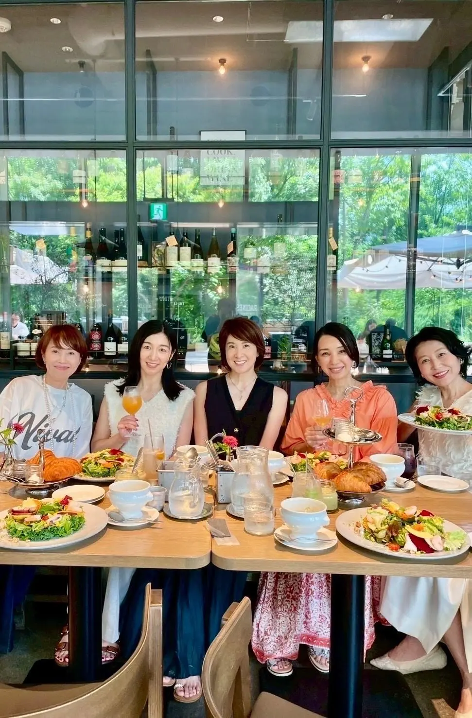 レストランで食事を楽しむ5人の女性。テーブルにはサラダ、パン、飲み物が並び、窓の外には緑豊かな景色が広がっている。