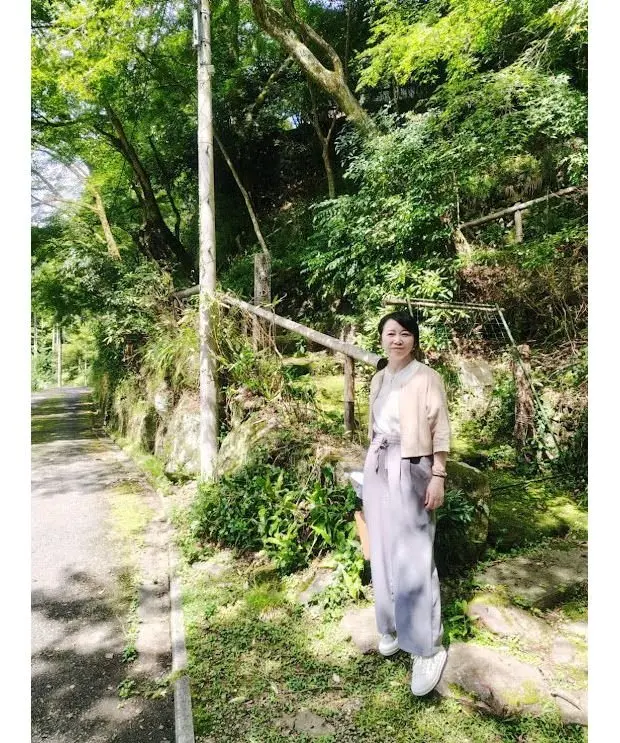 緑豊かな木々の中、道路脇に立つ女性。ベージュのトップスにグレーのワイドパンツ、白いスニーカーを着用。右手に茶色のバッグを持つ。