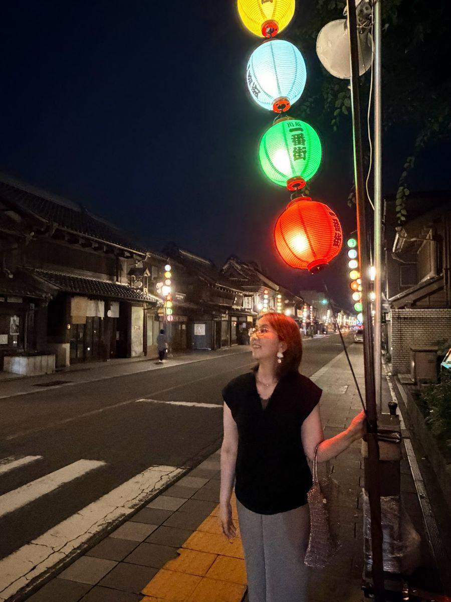 夜の街並みに立つ女性。赤、黄、緑の提灯が並び、背景には古い木造家屋が連なる。女性は黒いトップスとグレーのパンツを着用し、右手にバッグを持っている。