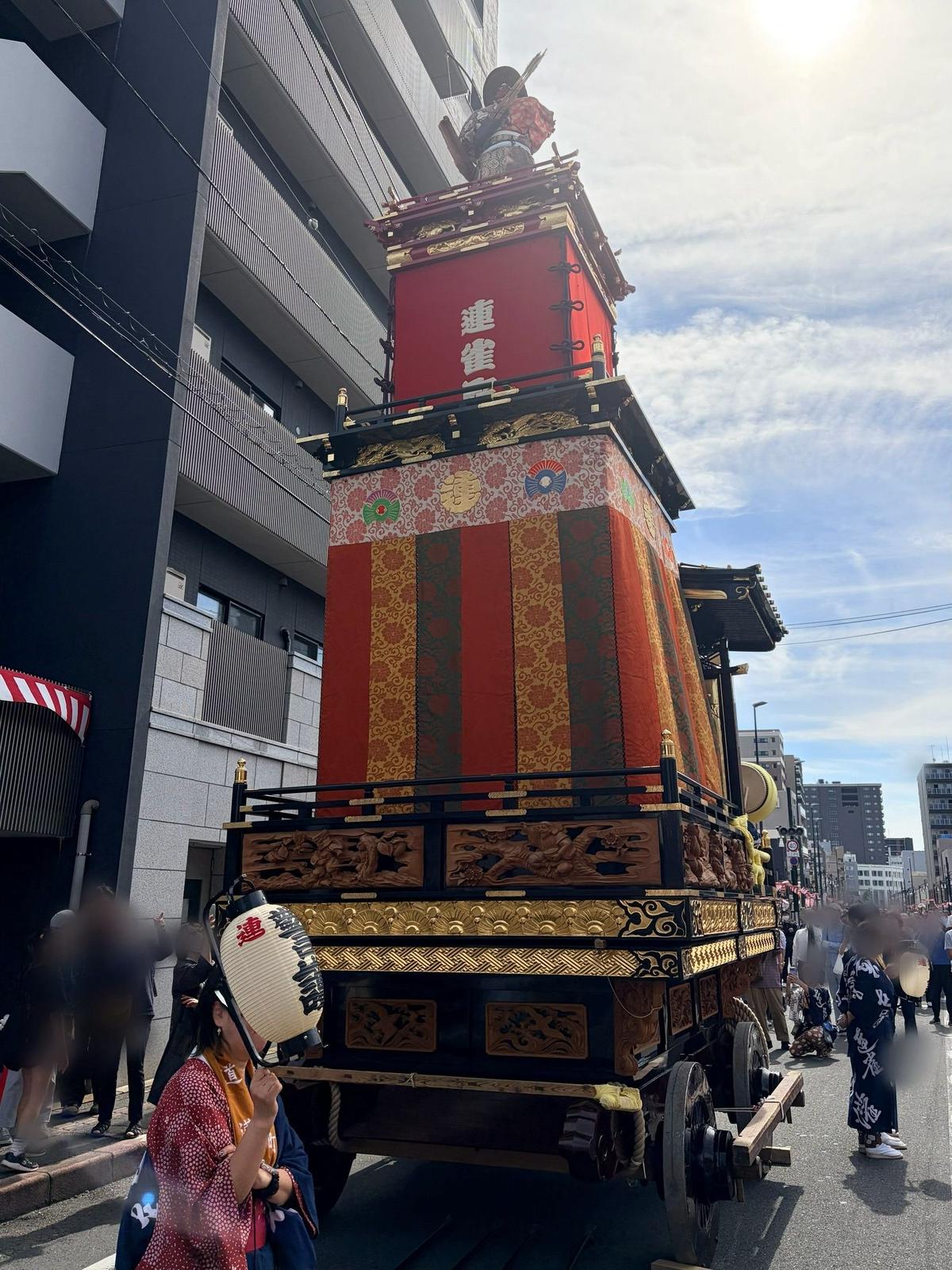 鮮やかな赤と金色の装飾が施された、日本の伝統的な山車が道路を移動している様子。山車の上部には武者人形が飾られ、周囲には見物客が見ている。