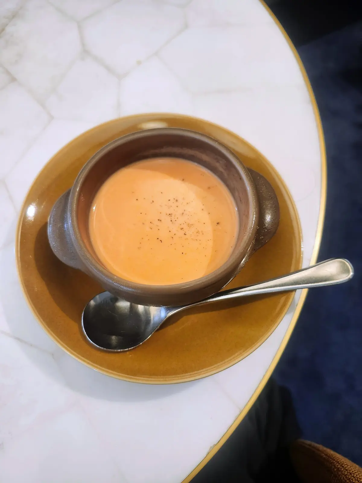 茶色の器に入ったオレンジ色のスープ。表面に黒胡椒が振りかけられている。黄色の皿に置かれ、銀色のスプーンが添えられている。白い六角形のテーブルの上。