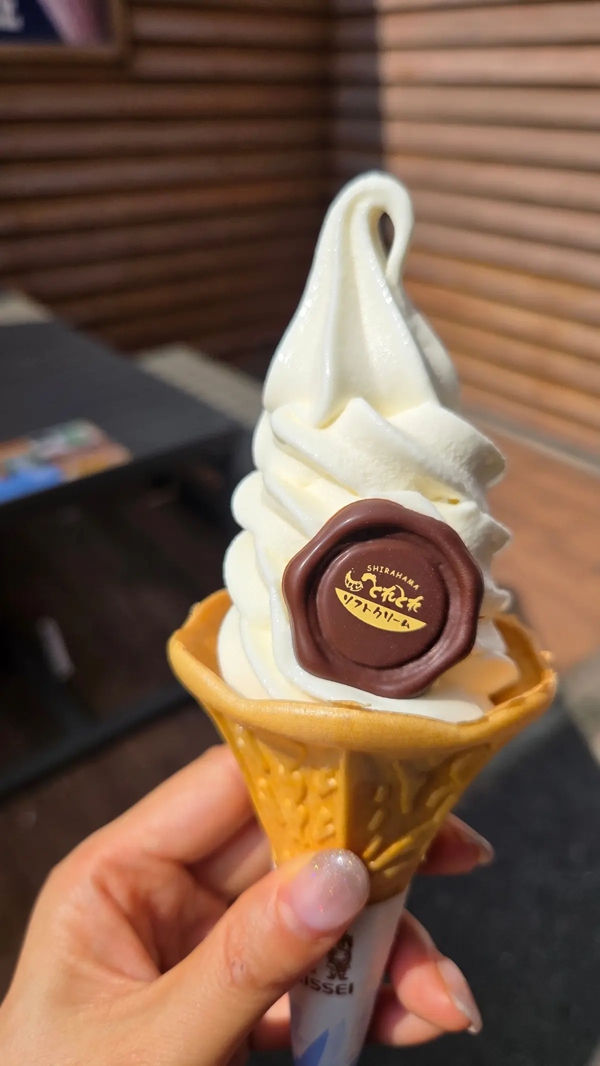 手で持たれたソフトクリーム。コーンに盛り付けられ、白いソフトクリームが螺旋状に高く積まれている。中央には「白浜ソフトクリーム」と書かれた丸いシールが貼られている。