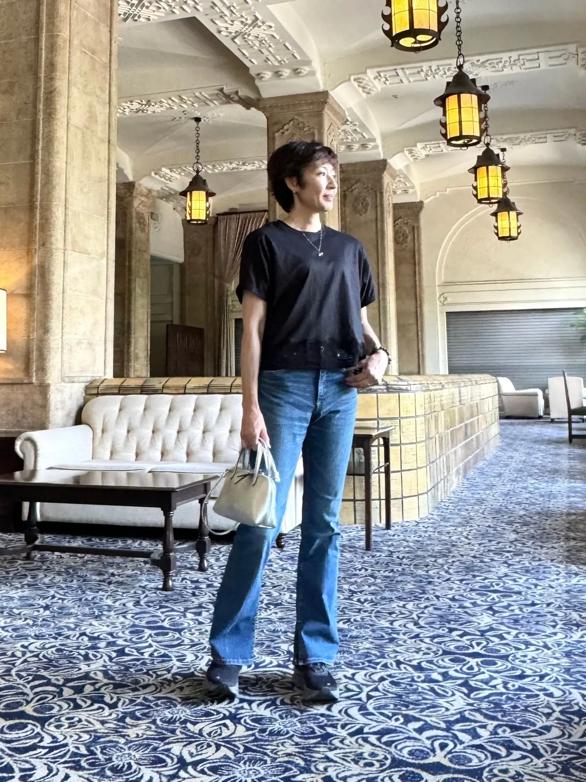 ホテルのロビーで、黒のTシャツにフレアジーンズを着用した女性が立っている。右手に白いハンドバッグを持ち、背景には装飾的な照明と絨毯が見える。
