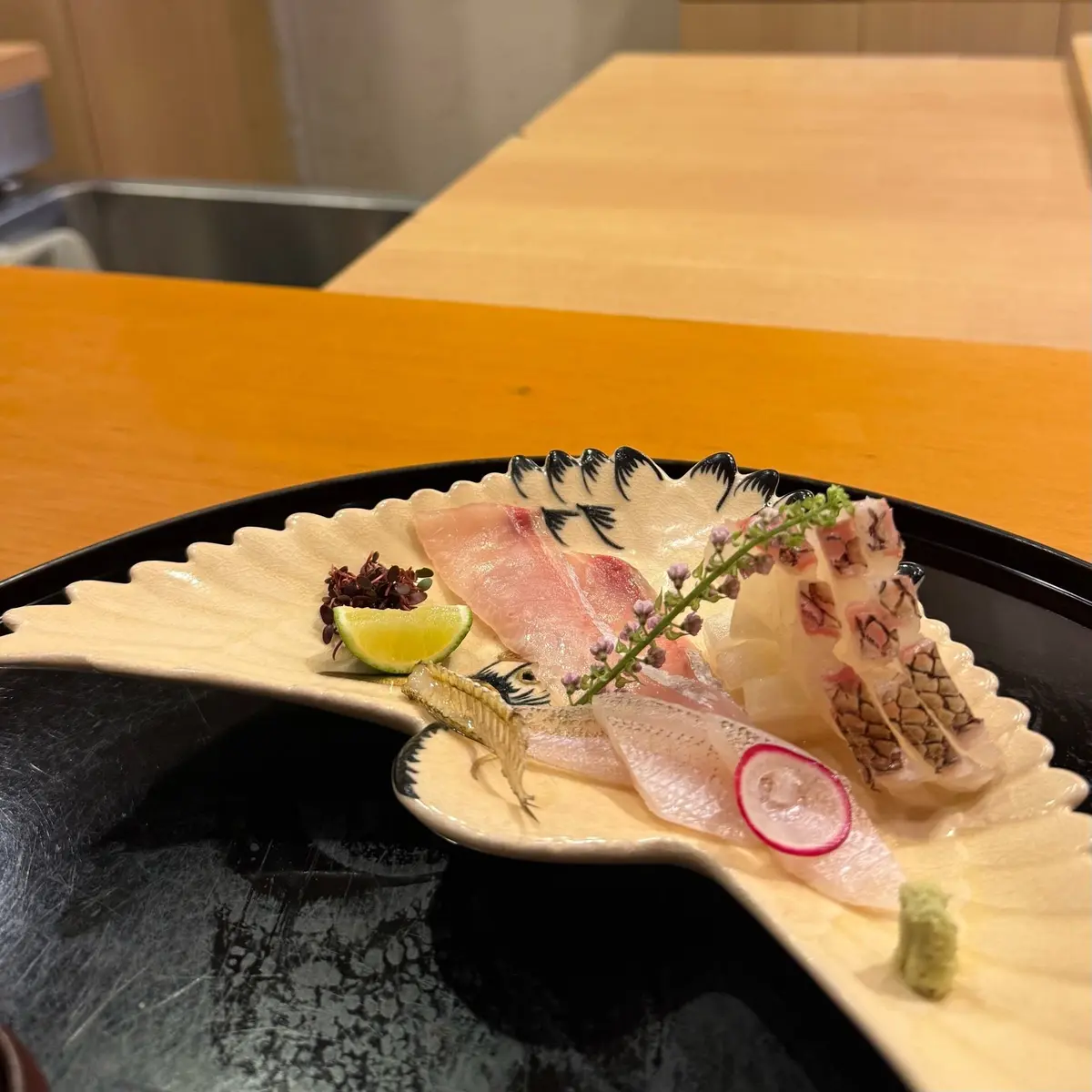 黒い漆塗りの皿に盛り付けられた、様々な種類の刺身。中央には白身魚、マグロ、光り物と思われる魚が並び、付け合わせとして赤紫色の海苔、ライム、ミョウガ、ワサビが添えられている。