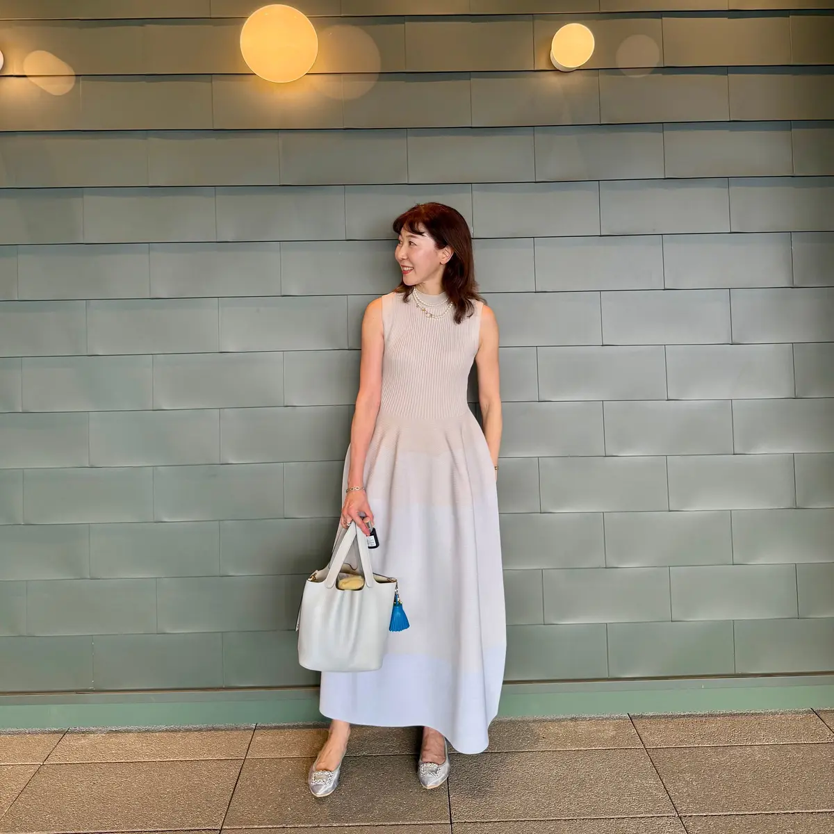 白いAラインのワンピースを着た女性が、緑色の壁の前で微笑んでいる。右手に白いハンドバッグを持ち、足元にはシルバーの靴。背景には白い球体型の照明が並んでいる。