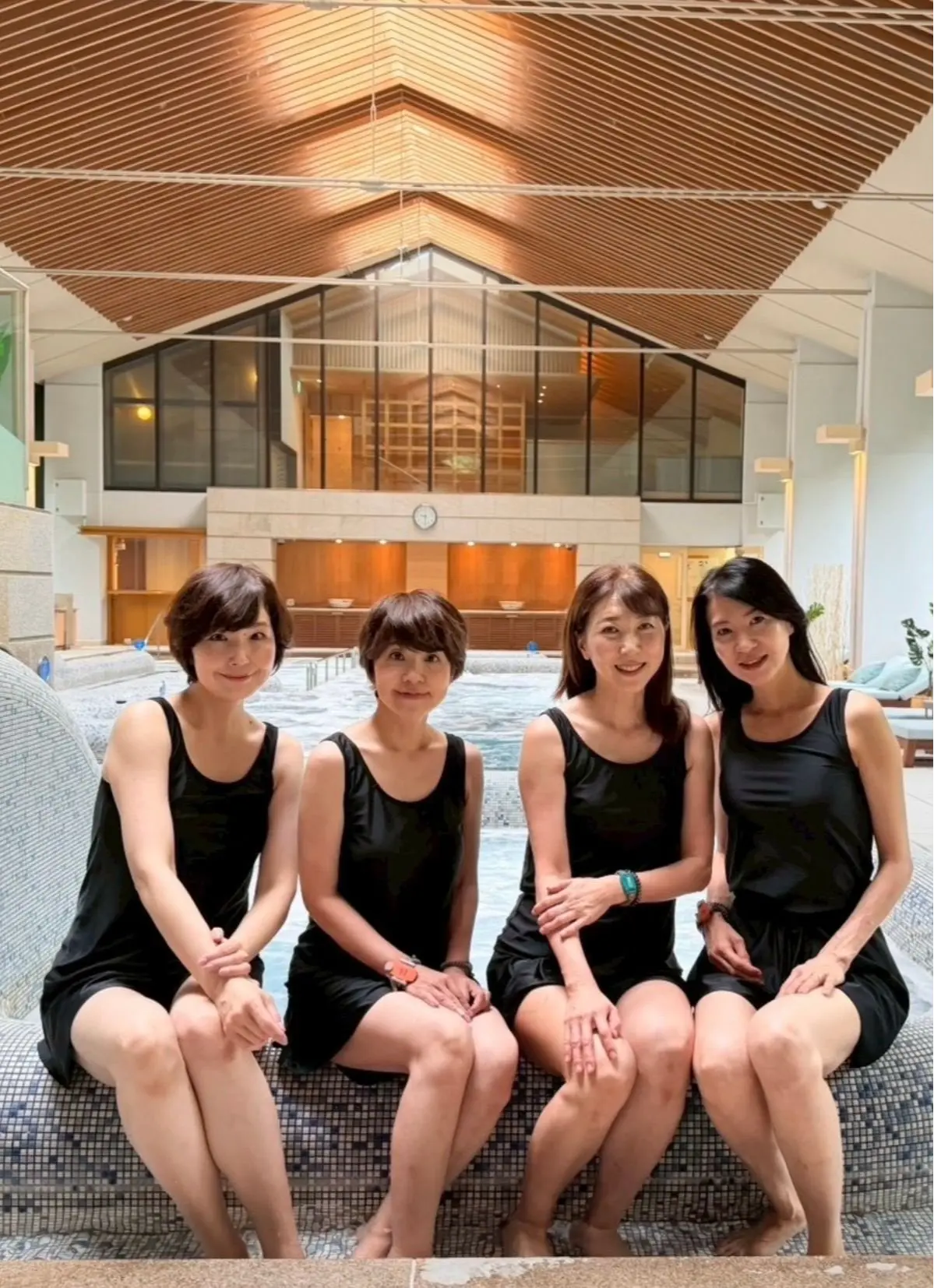 4人の女性が黒い水着でプールサイドの椅子に並んで座り、プールとモダンな建物が見える背景。