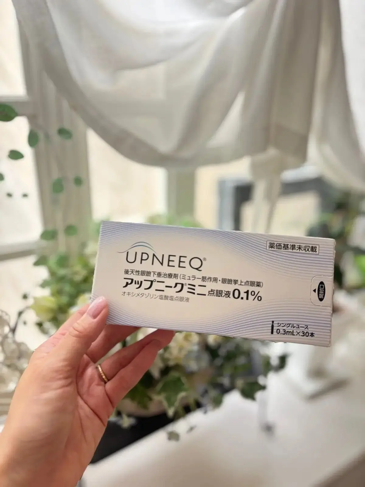 UPNEEQの医療用点眼剤パッケージを女性の手が持つ。白い背景に緑の葉が散らばり、窓の外の建物がぼやけて見える。