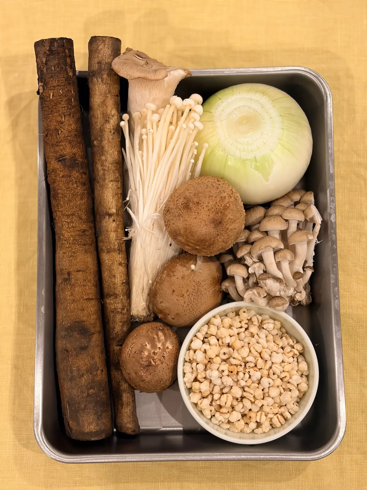 金属製のトレイに入った、キノコ、根菜、穀物の食材セット。長めのレンコン、椎茸、舞茸、球状の洋ナシのようなキノコ、そして白玉のような穀物が並んでいる。