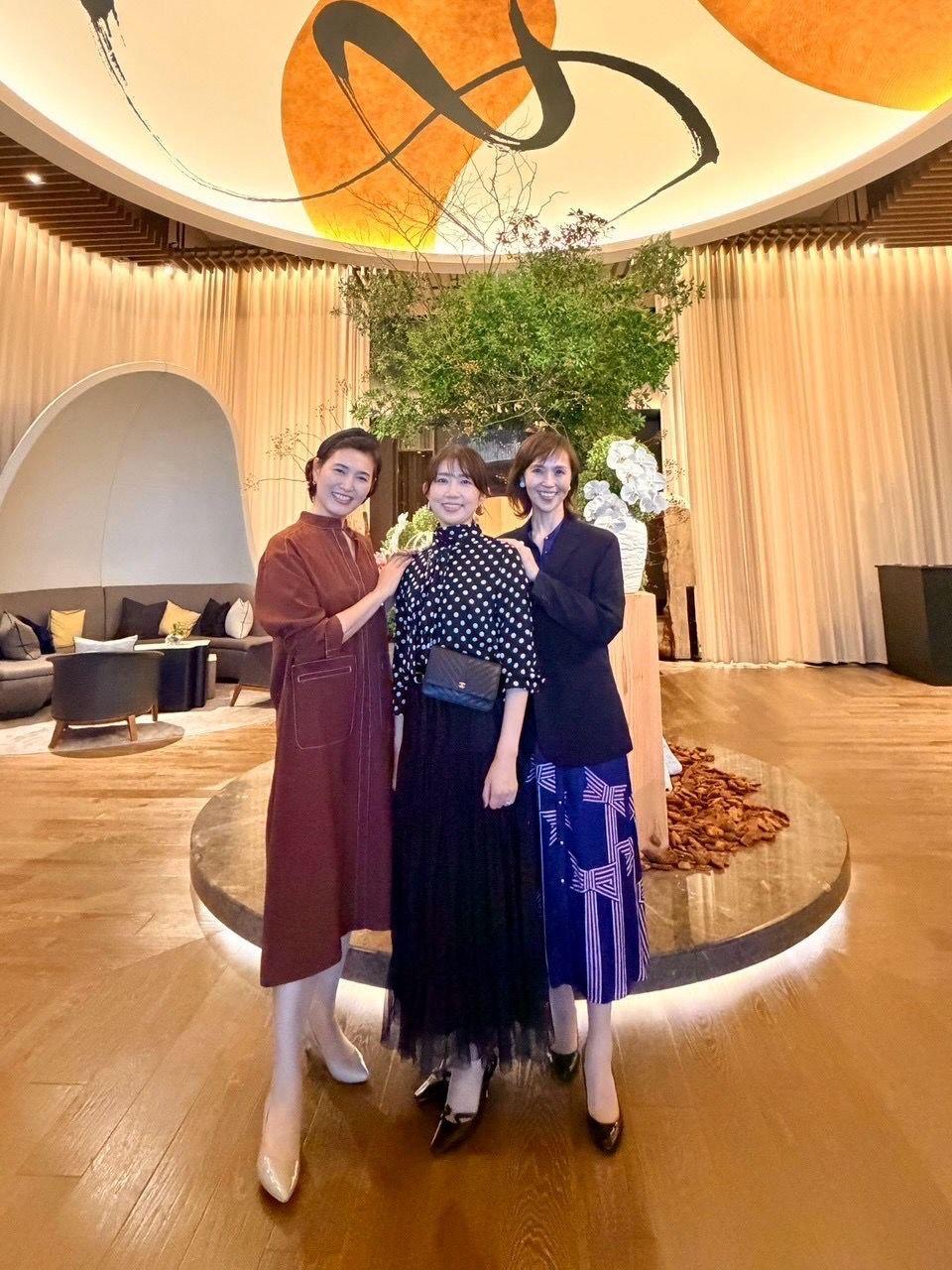 豪華な内装の空間で、3人の女性が笑顔で並んで立っている。左から茶色のワンピース、黒のドット柄トップスに紺のスカート、黒のジャケットに紺のスカートを着用。背景にはオレンジ色の円と植物の装飾がある。
