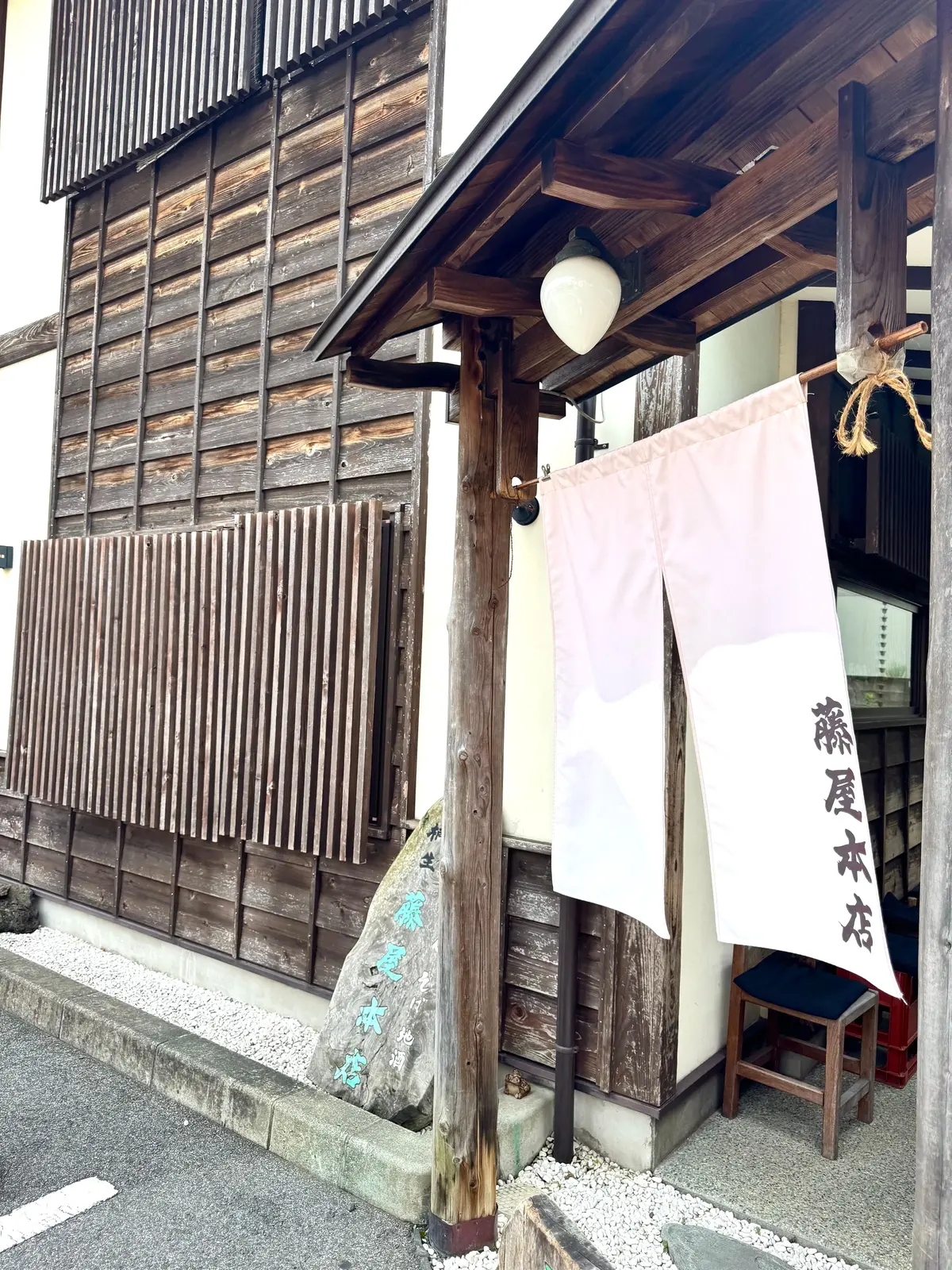 木造の建物の入り口。木製の格子戸が特徴的で、白い幕と看板が掲げられている。看板には日本語で「藤屋木村」と書かれている。照明が吊り下げられ、石畳の道が続く。