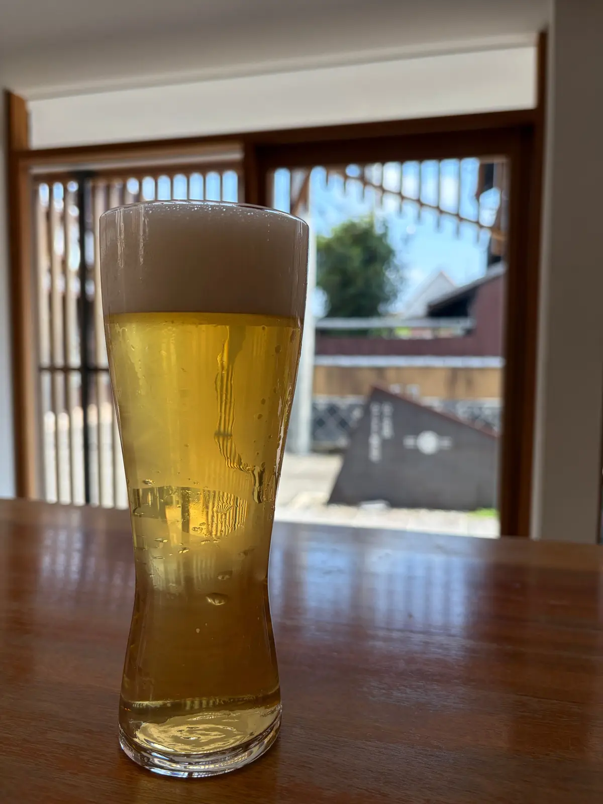 木製のテーブルに置かれた、泡が乗った麦わら色のビールグラス。背景には木製の格子戸と、庭の風景が見える。