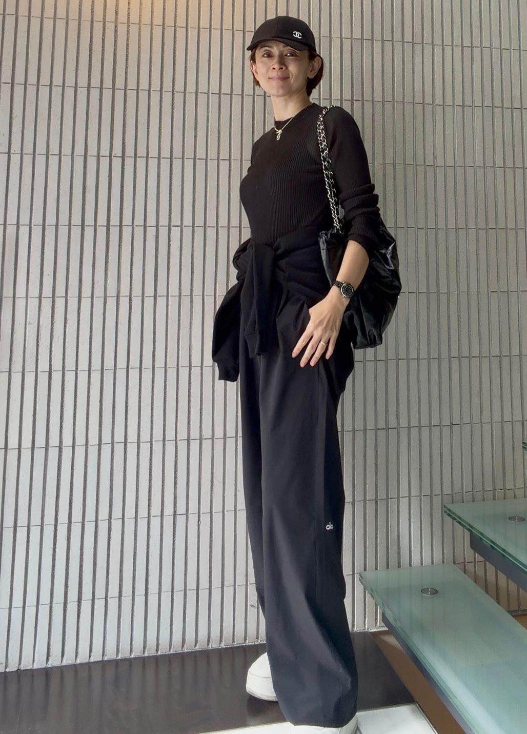 黒いキャップを被った女性が、黒いニット、スウェットパンツ、パーカーを着用して立っている全身写真。右手に黒いバッグを持ち、左手首には腕時計をしている。背景はグレーのタイル壁。