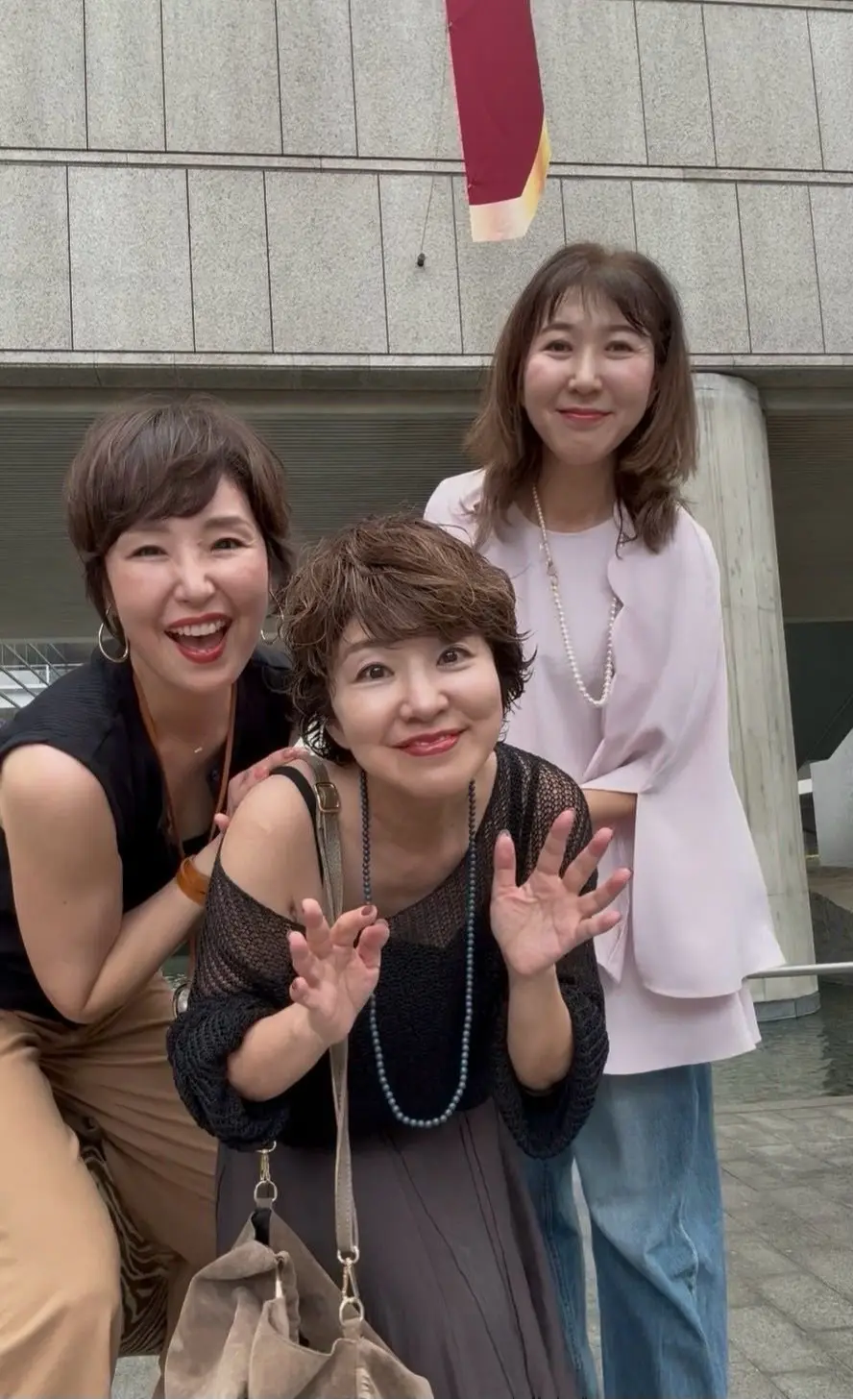 3人の女性が屋外で笑顔を見せている写真。中央の女性は黒いオフショルダーのトップスとスカートを着用し、手を広げてポーズをとっている。左と右の女性はそれぞれベージュのトップスと白いシャツを着ている。背景にはグレーの建物と、赤と白の旗が見える。
