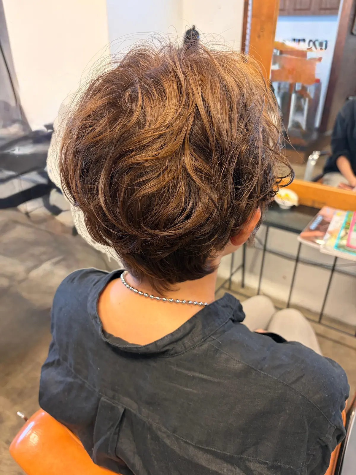 茶色の縮れ毛の女性のバックショット。首元には銀色のシンプルなネックレス。後ろ髪はふんわりとボリュームがあり、襟足は短くカットされている。