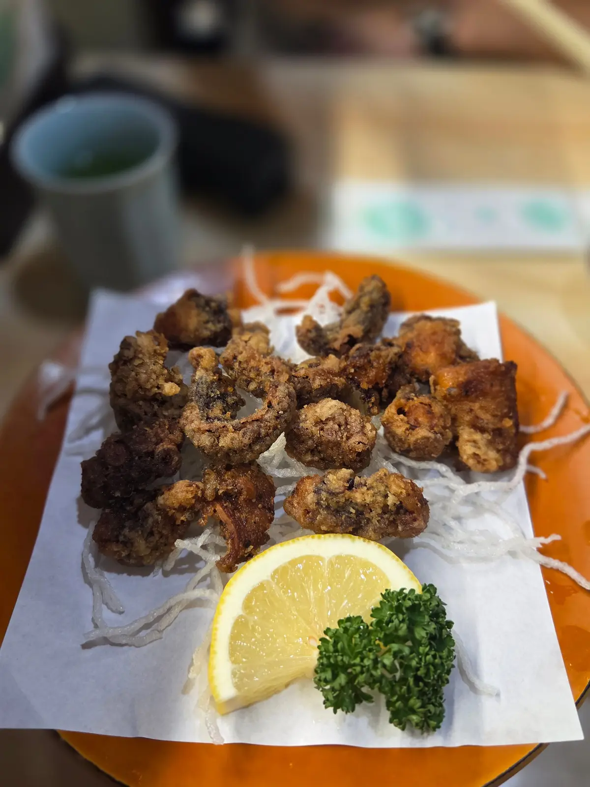 オレンジ色の皿に盛り付けられた、鶏の心臓の唐揚げ。白髪のネギとレモンスライス、パセリが添えられている。背景には飲み物と箸が見える。