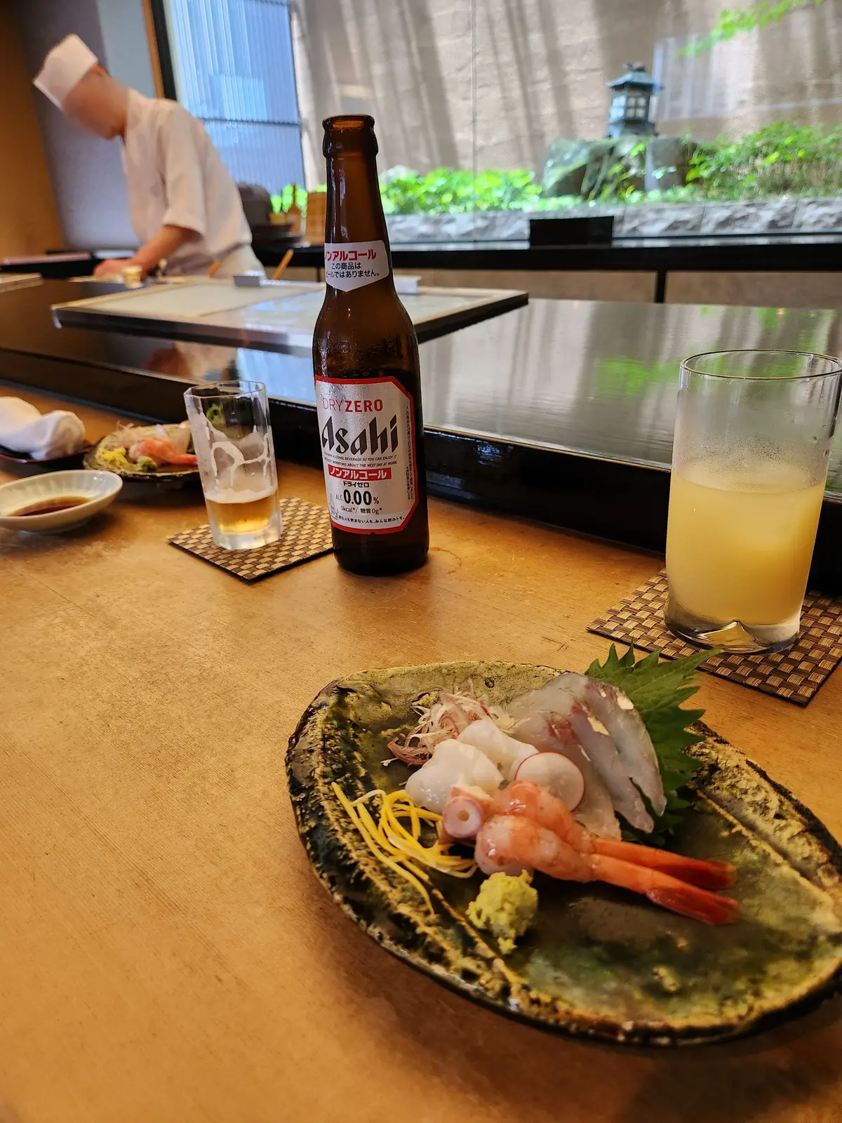 木製テーブルに、海鮮料理の盛り合わせ、ビール瓶、グラスが置かれている。背景にシェフが作業している。
