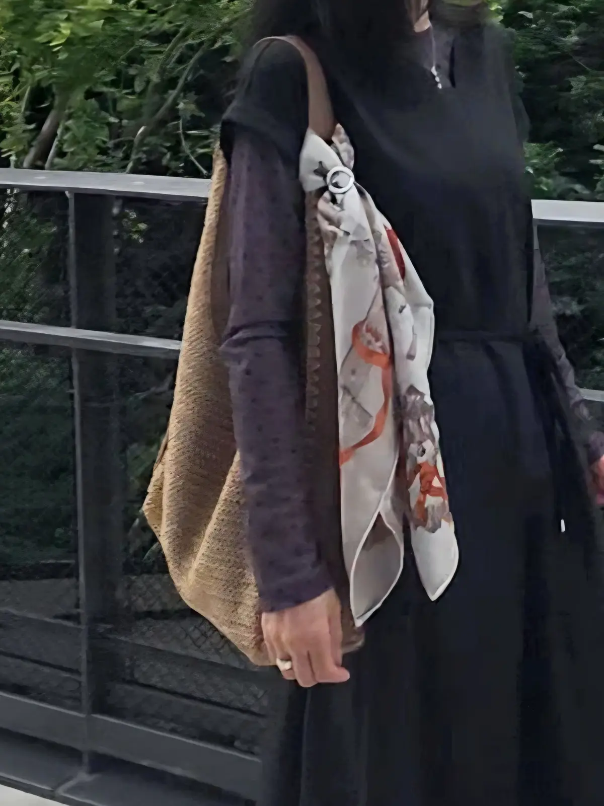 黒の服を着た女性が、赤と白の模様のあるスカーフを首に巻いています。手には茶色のショルダーバッグをかけ、屋外の緑豊かな背景が広がっています。