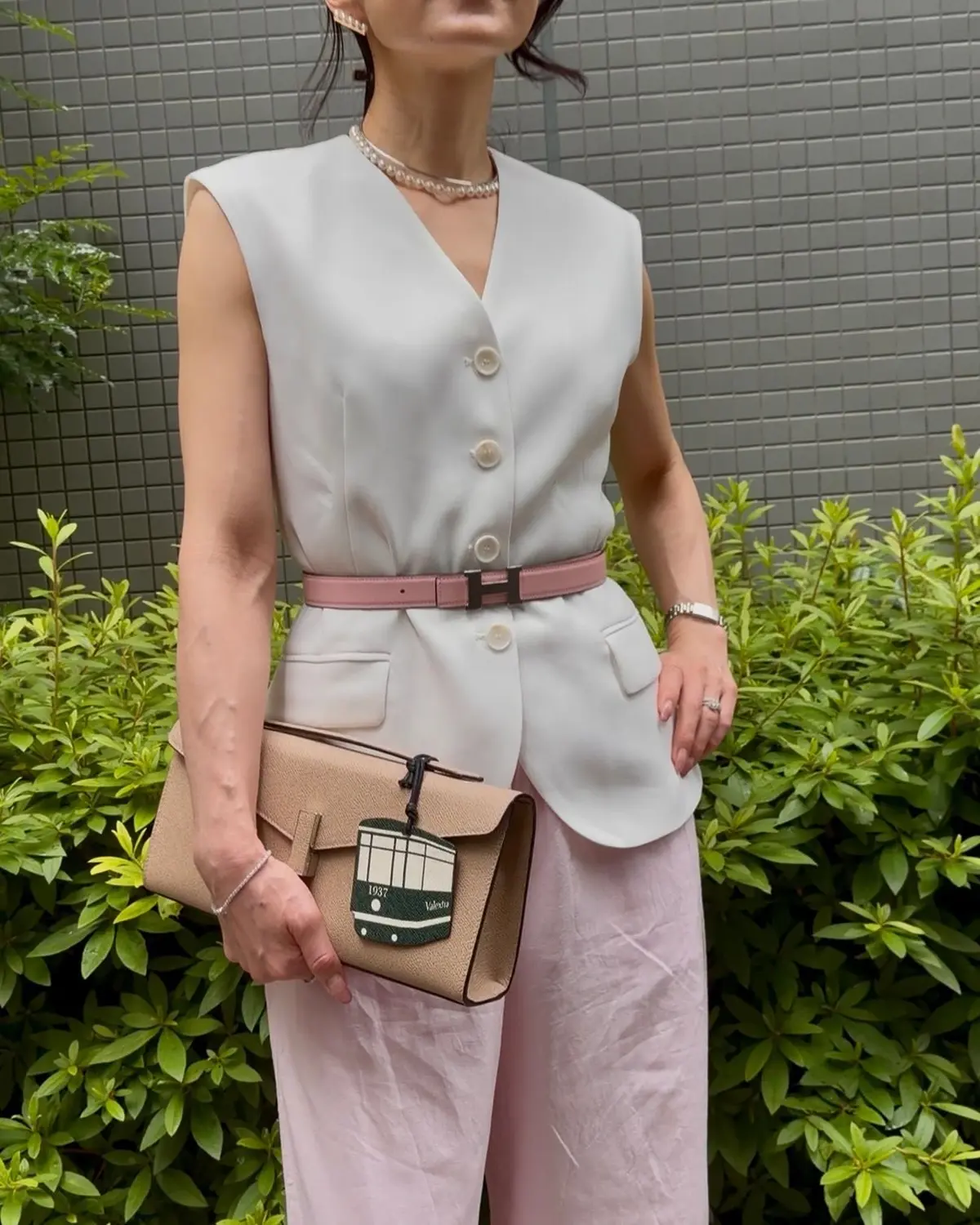 白のノースリーブのトップスにピンクのベルトを締め、ピンクのスカートを着た女性が、ベージュのクラッチバッグを持っています。背景には緑の低木が並んでいます。