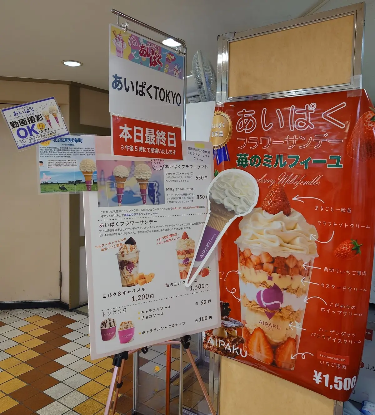 「あいぱく」の屋台の様子。赤と白の屋台には、ソフトクリームやドリンク、お菓子のメニューが掲示されている。左には「動物園OK!」の案内、右には「フラワークッキー」の看板が立っている。背景には、砂浜と青い空が広がり、夏らしい雰囲気を醸し出している。