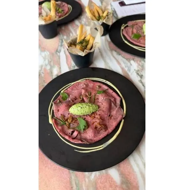 黒い皿に盛り付けられた薄切り牛肉、緑色のソース、香草、黄色の線状ソース。背景にはフライドポテトと他の料理が写っている。