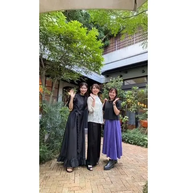 緑豊かな庭園を背景に、3人の女性が立っている。左は黒いロングドレス、中央は白いレースのトップスと黒いパンツ、右は黒いトップスに紫色のスカートを着用。それぞれ笑顔で手を振っている。