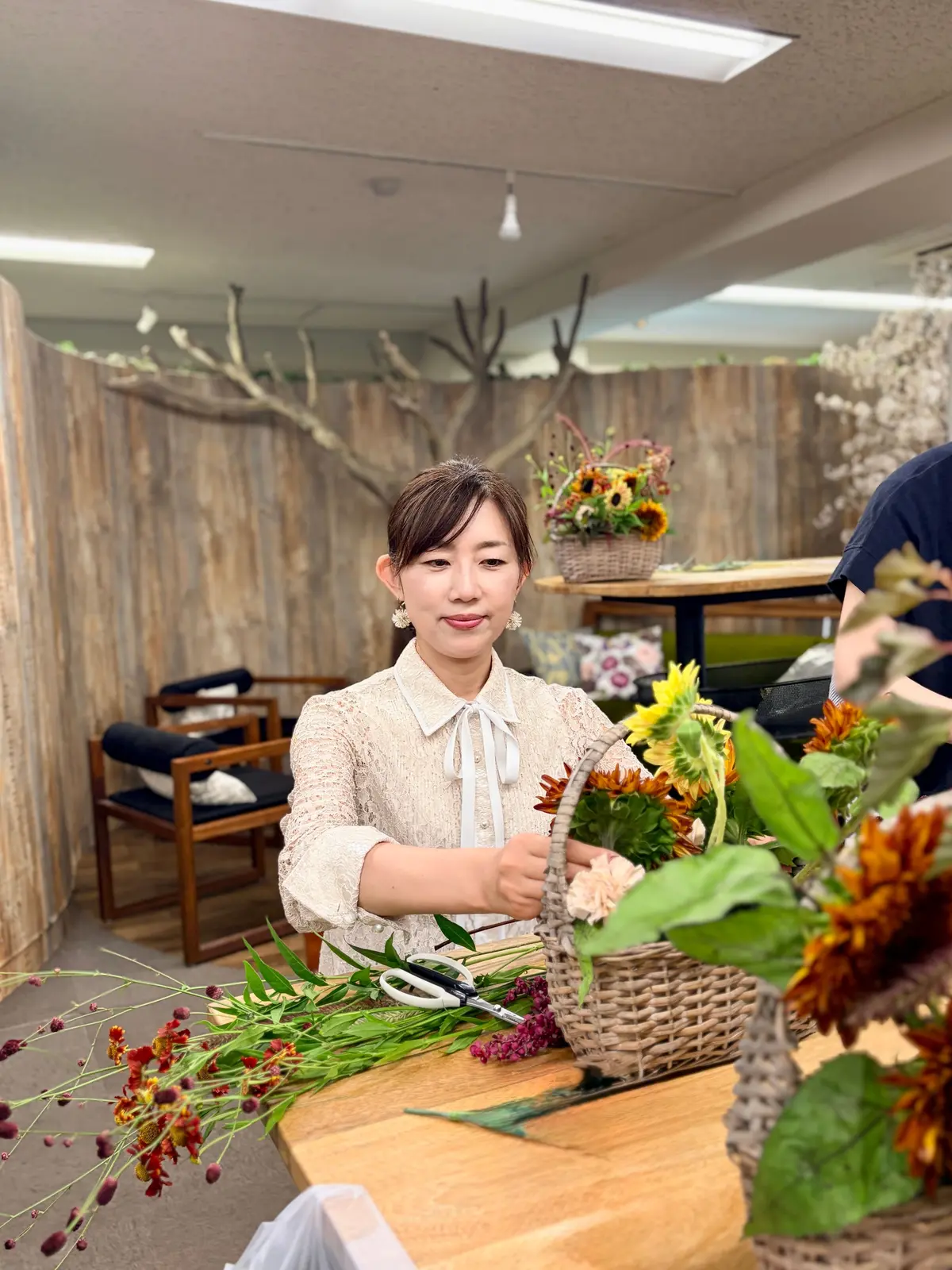 笑顔の女性が、花を編み込む籠を手に作業している。木製のテーブルには花束や剪定バサミが置かれ、背景には木製の壁と装飾的な木の枝がある。