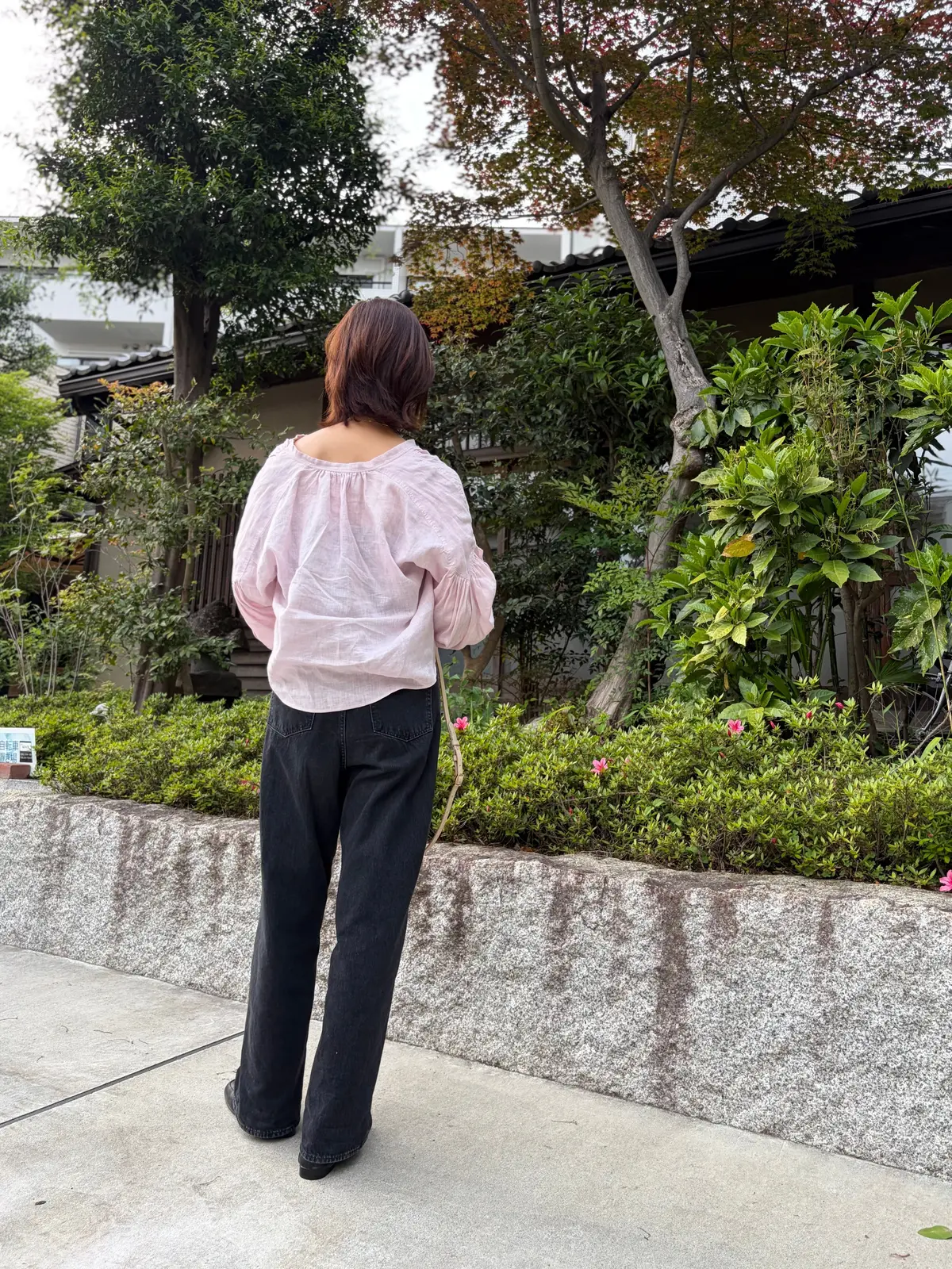 薄いピンクのプルオーバーとデニムパンツを着た女性が、背面から見え、緑豊かな庭園を背景に歩いています。
