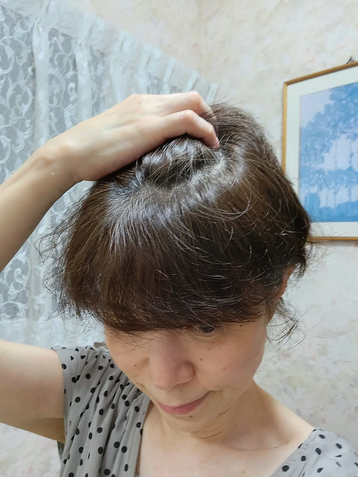 女性が手で頭を掻いている様子。茶色の髪に白い部分が見え、薄毛の兆候がある。背景にはカーテンと絵画が掛かっている。