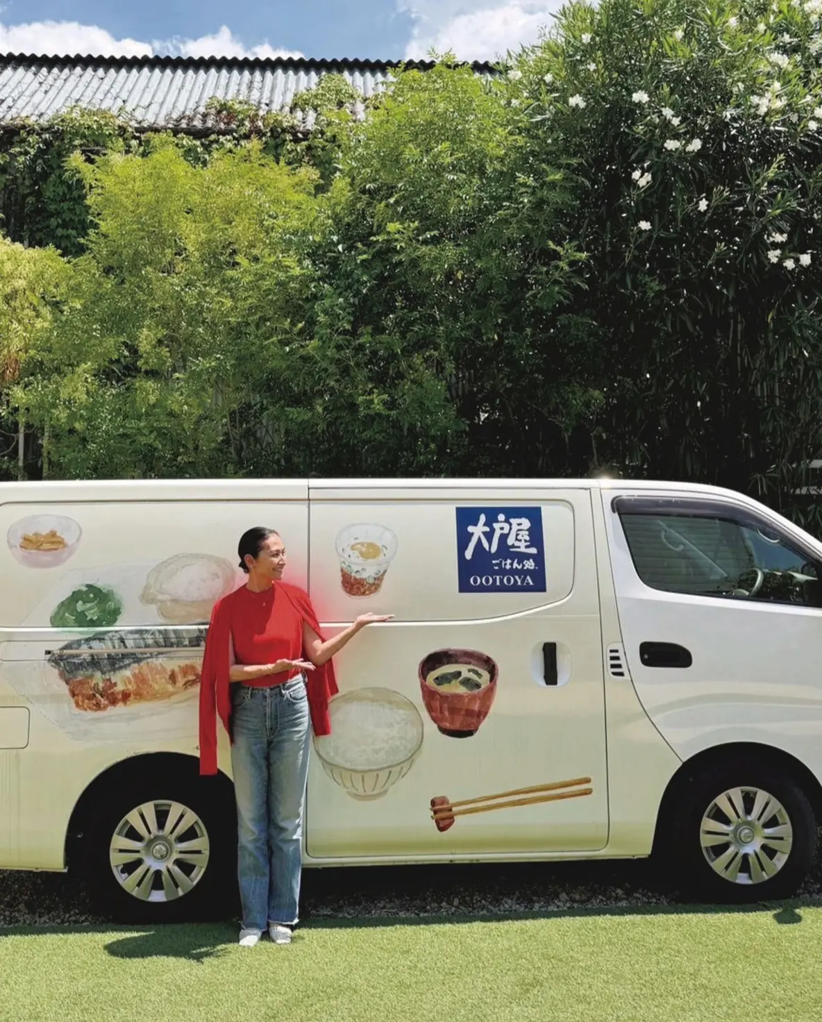 緑豊かな背景をバックに、白の移動販売車が置かれている。車体には「大戸屋」のロゴと、料理のイラストが描かれている。赤いシャツにデニムを着用した女性が、車に寄り添って立っている。