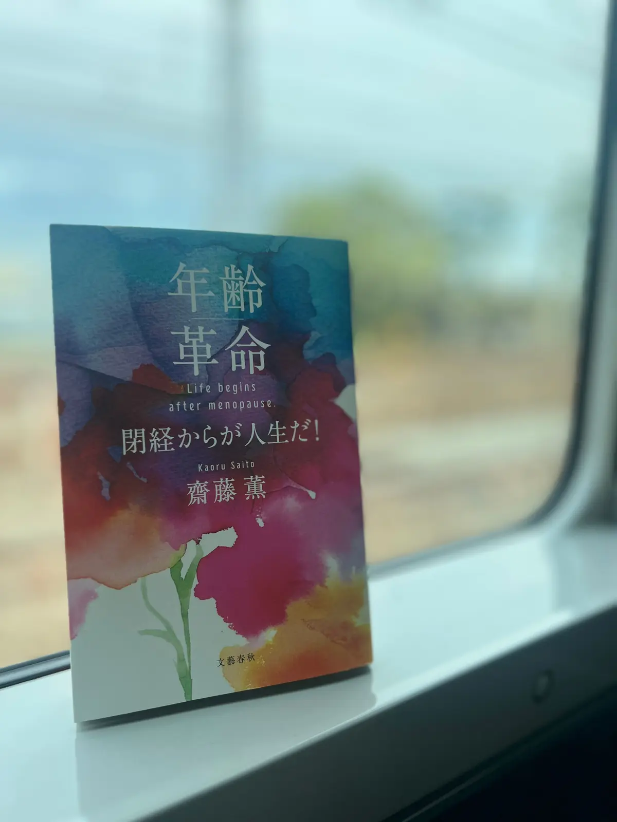 水彩風の表紙に「年齢革命 Life begins after menopause. 閉経からが人生だ!」と書かれた本が窓辺に置かれている