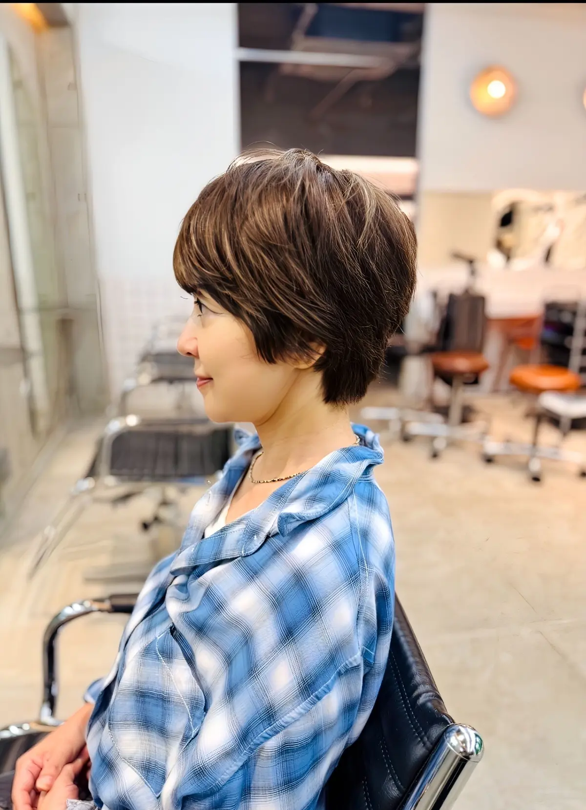 青いチェック柄シャツの女性が、美容室の椅子に座り、短い髪を整えている。背景には鏡と椅子が見える。