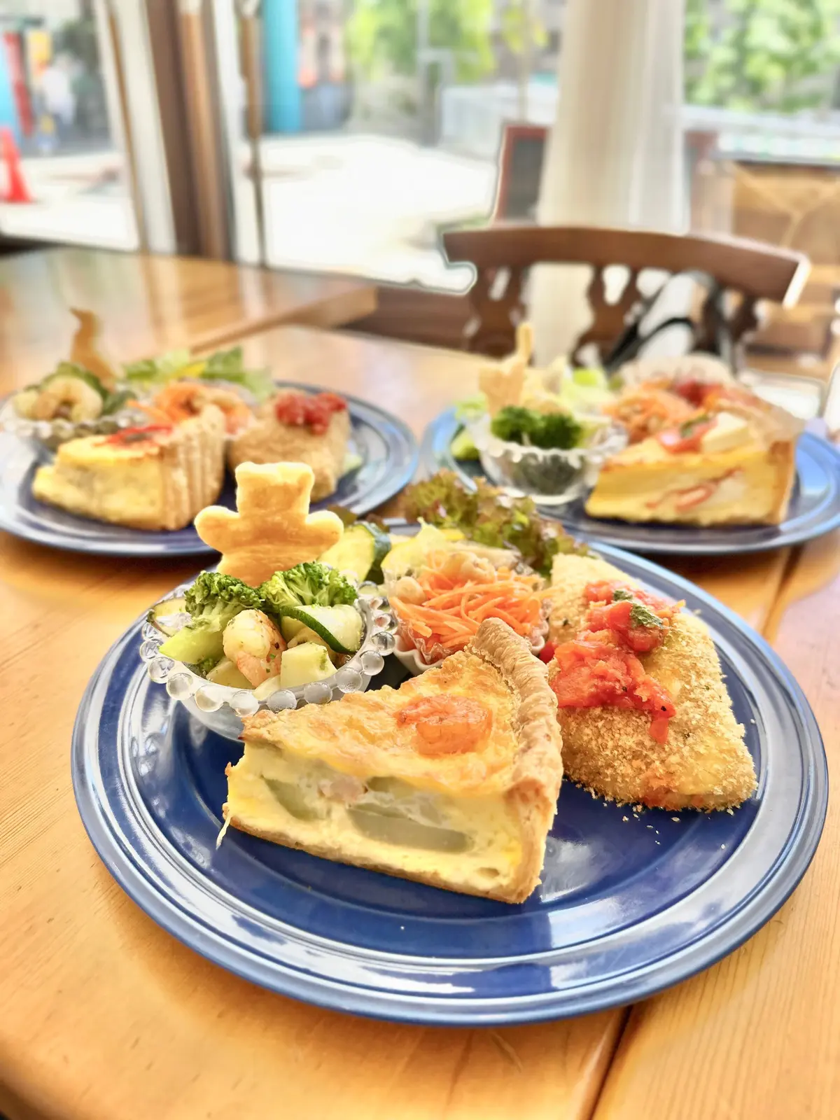 木製のテーブルに、青いプレートに盛りられた色とりどりの料理が並ぶ。中央には野菜とエビのサラダ、トマトをトッピングしたキッシュ、揚げたエビの料理が置かれている。