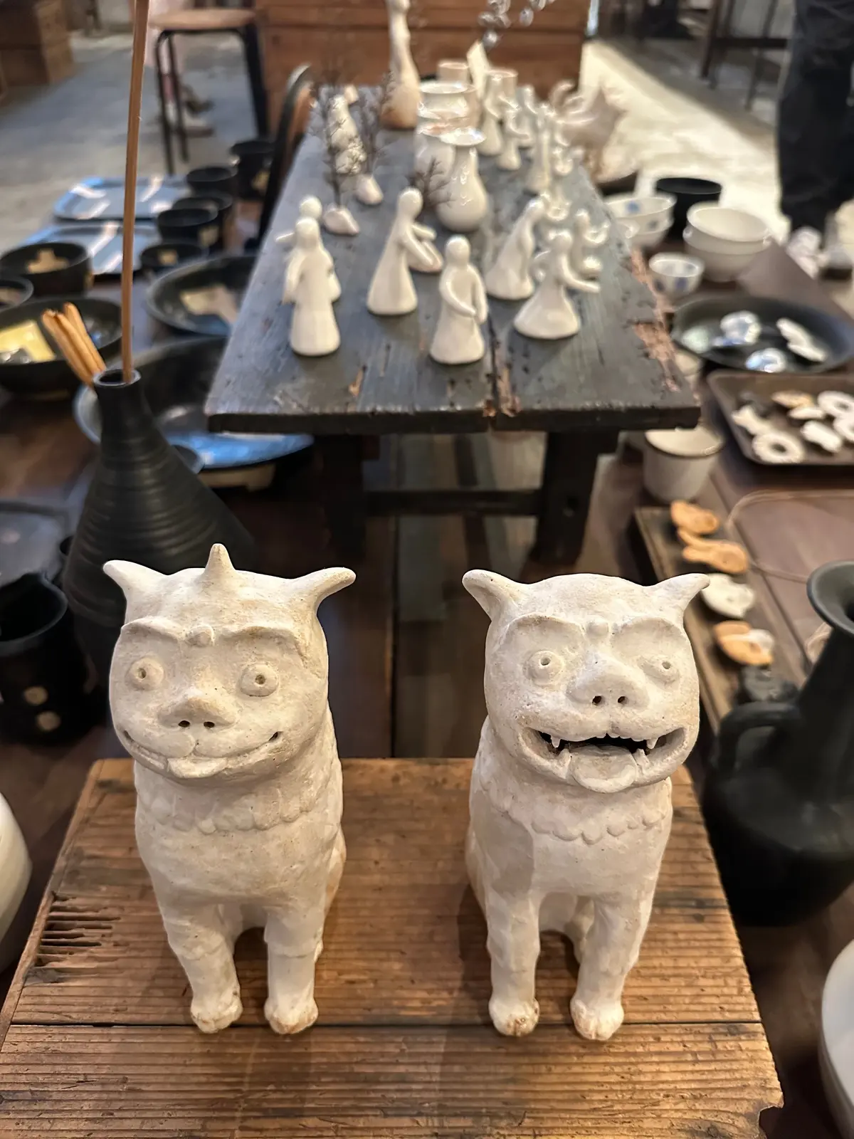 白い陶器の猫像二体、木製テーブルに並ぶ陶器の食器や人形、背景に黒い壺が置かれた陶器の展示場