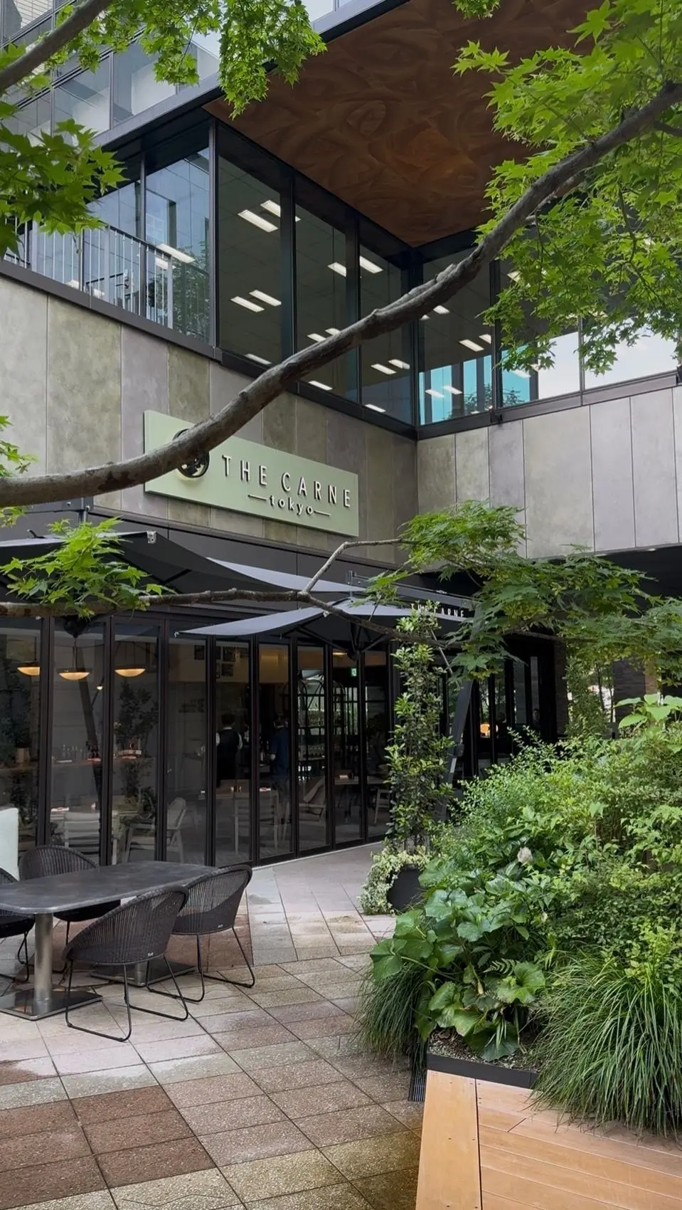 THE CARNE TOKYOの屋外テラス、木製の天井とガラスの壁、緑の植物とテーブルと椅子が配置されたモダンな雰囲気