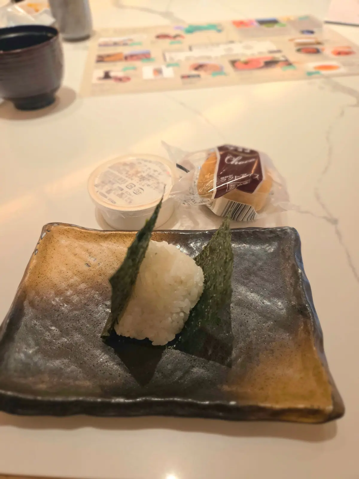 茶色い陶器の皿に、海苔で巻かれた白いご飯が2つ置かれている。皿の隣には、白い容器とチョコレートの個包装が置かれている。背景には、黒い湯呑みとメニューが写っている。