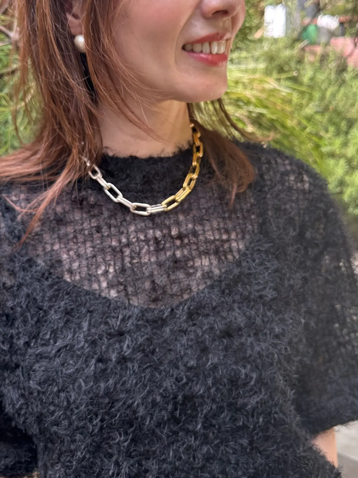 ふわふわの黒いフェイクファースウェーターを着た女性で、右耳に真珠のイヤリング、首に金色のチェーンネックレスを身につけています。背景は緑の植物です。