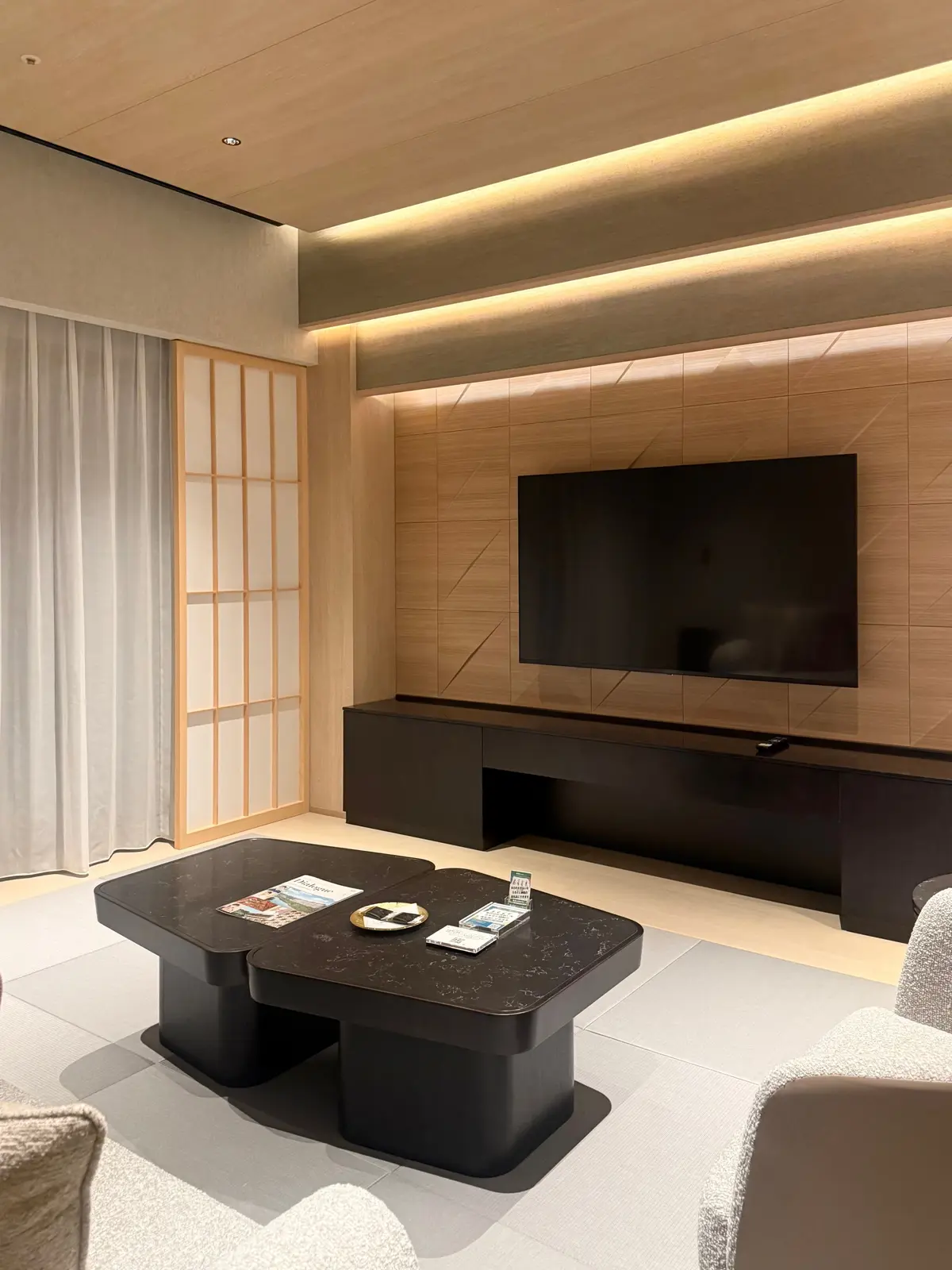 モダンなリビングルームで、木製のテレビボードに大型スクリーンが設置され、黒のテーブルには雑誌や小物があり、和風の障子とカーテンが備わっている
