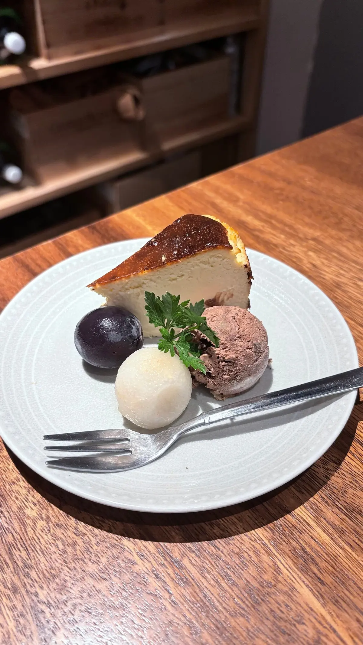 グレーの皿に、チーズケーキの切り口、プラム、シャーベット、パセリが盛り付けられている。フォークが添えられている。