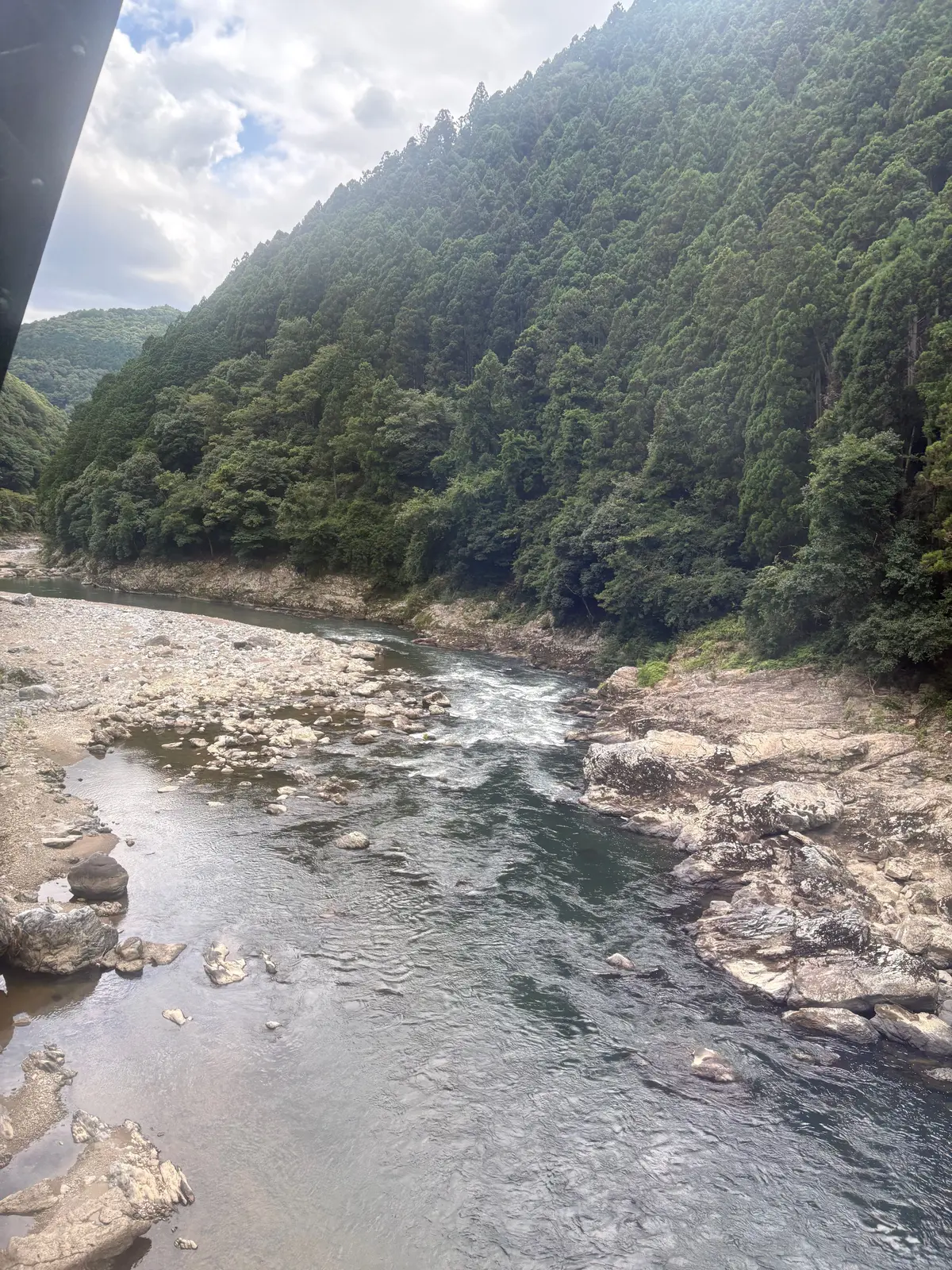 緑豊かな山々と岩場が広がる川の風景。水は流れが速く、川底には大小の岩が点在している。空は曇り、明るい光が差し込んでいる。
