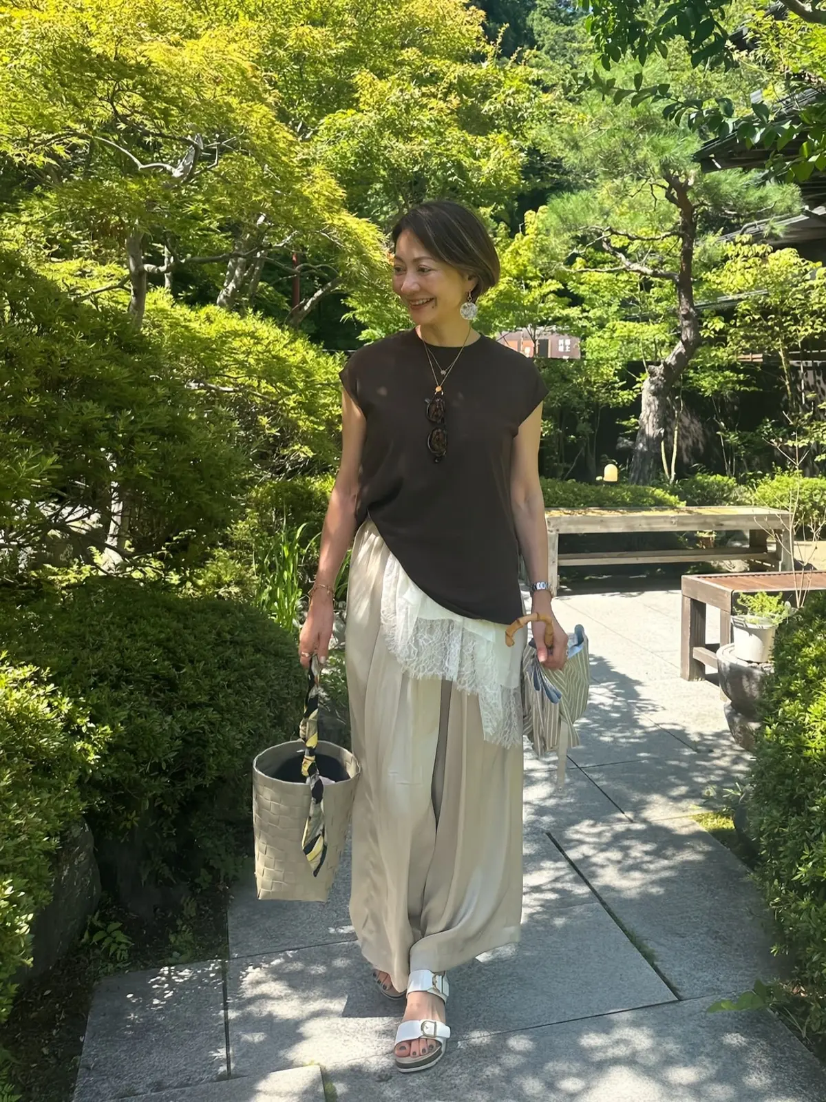 茶色のタンクトップに、ベージュのドレープパンツとレースのインナーを合わせた女性。白いサンダルを履き、黄色のバッグとサングラスを持つ。背景には緑豊かな庭園。