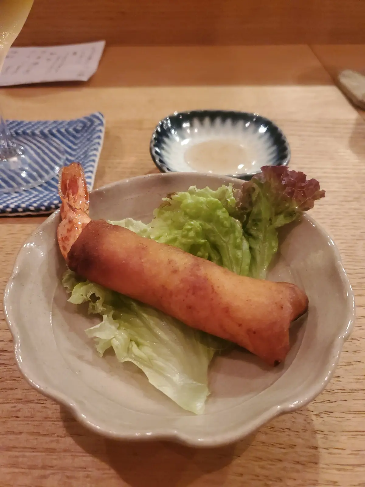木製のテーブルに置かれた小皿に、エビの春巻きとレタスが盛り付けられている。背景には、ソースが入った小皿とランチョンマットが写る。
