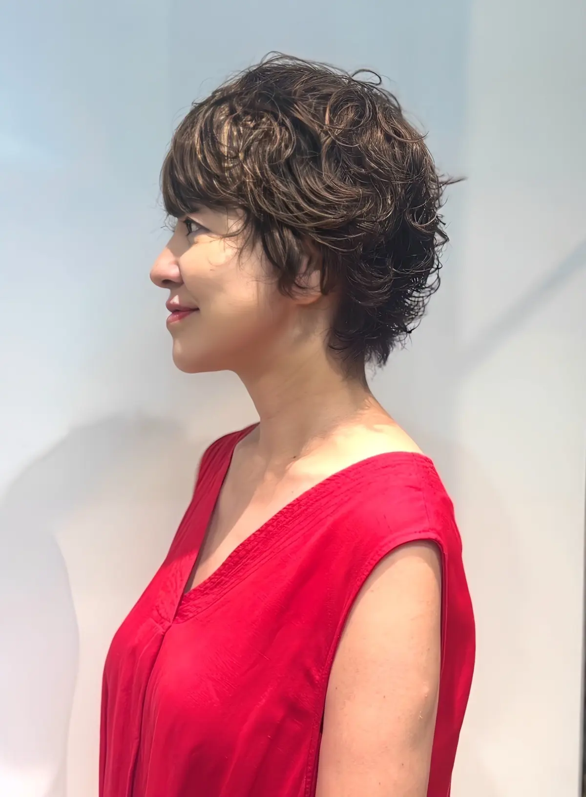 赤いVネックのワンピースを着た女性の横顔。肩上りの濃い茶色の縮髪で、右側に光が当たっている。背景は白い壁と窓。