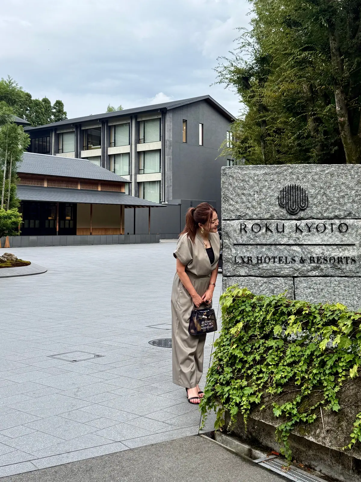 京都のホテル、ROKU KYOTOの入り口に立つ女性。背景にはモダンなホテルと緑豊かな植栽が見える。