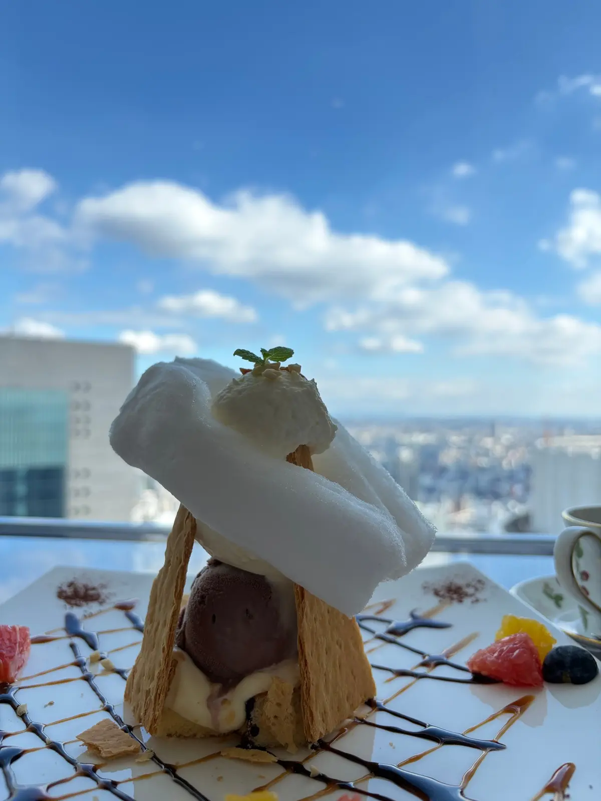 チョコレートアイスクリームとメレンゲが乗ったデザート。ワッフルの器に盛り付けられ、フルーツやソース、クッキーなどが添えられている。背景には高層ビル群と青空。