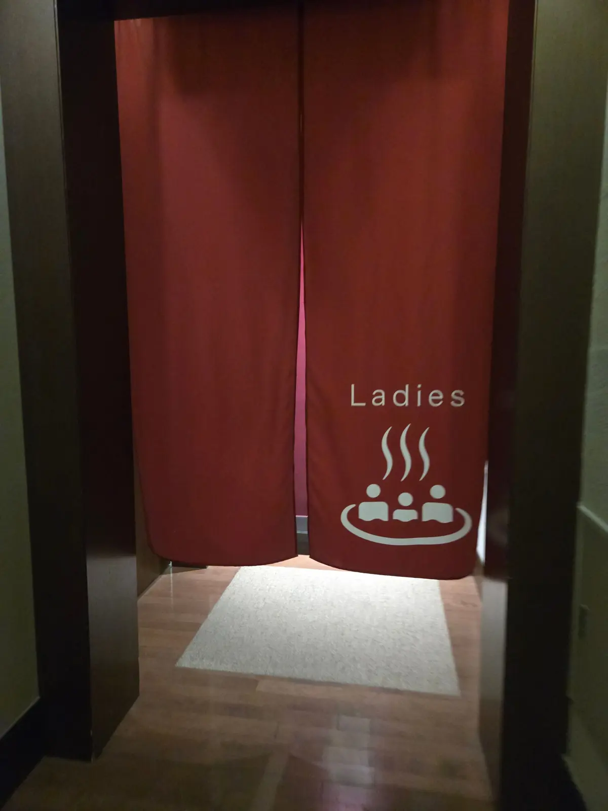 赤色のカーテンで仕切られた女性用トイレの入り口。ドアには「Ladies」の文字と、湯気と人影のイラストが白で描かれている。床には小さなマットが置かれている。